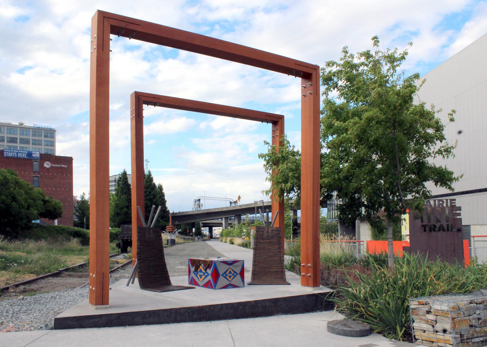 The completed art piece, with two chairs under a large wooden frame next to a number of Native American trading and luxury goods 