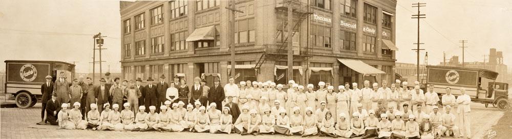 Photo of the employees of the Tacoma Biscuit and Candy Co. in 1921 Tacoma, WA who are posed in rows in front of the companys building.